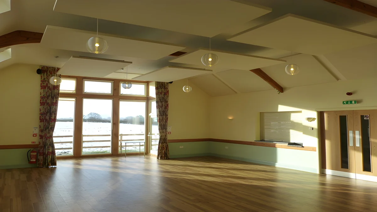 Suspended acoustic panels and pendant lights in Ryton Village Hall, looking towards its full-height windows.