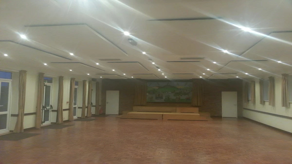 Rows of white acoustic ceiling panels and downlights above the open wooden floor of Radnage Village Hall, looking towards its stage.
