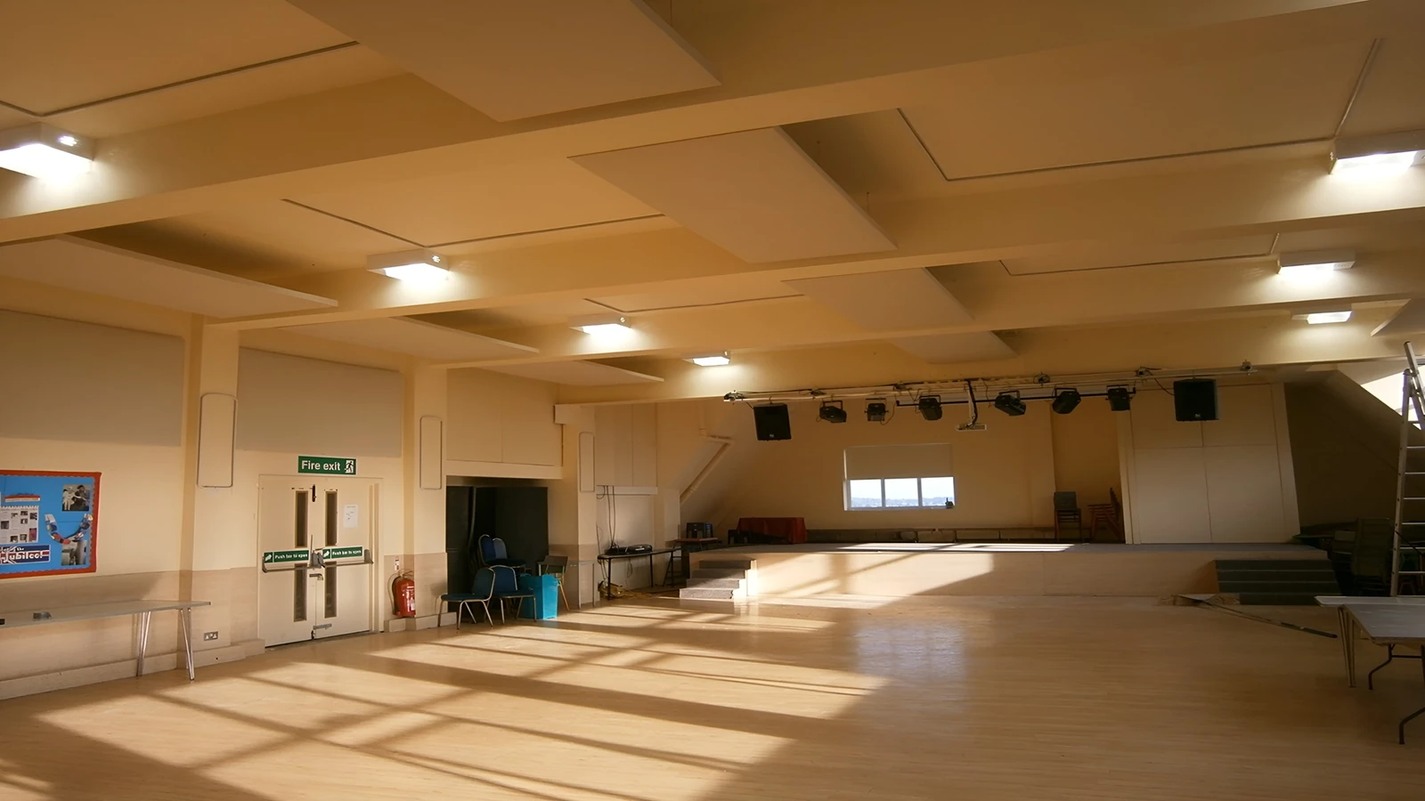 Large pale suspended ceiling panels above the wooden floor and stage of Nancy Reuben Primary School hall.