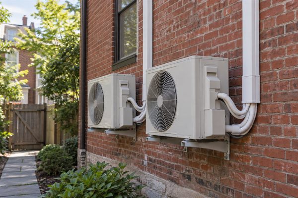 Two heat pump outdoor units on wall brackets on the brick side of a rowhouse
