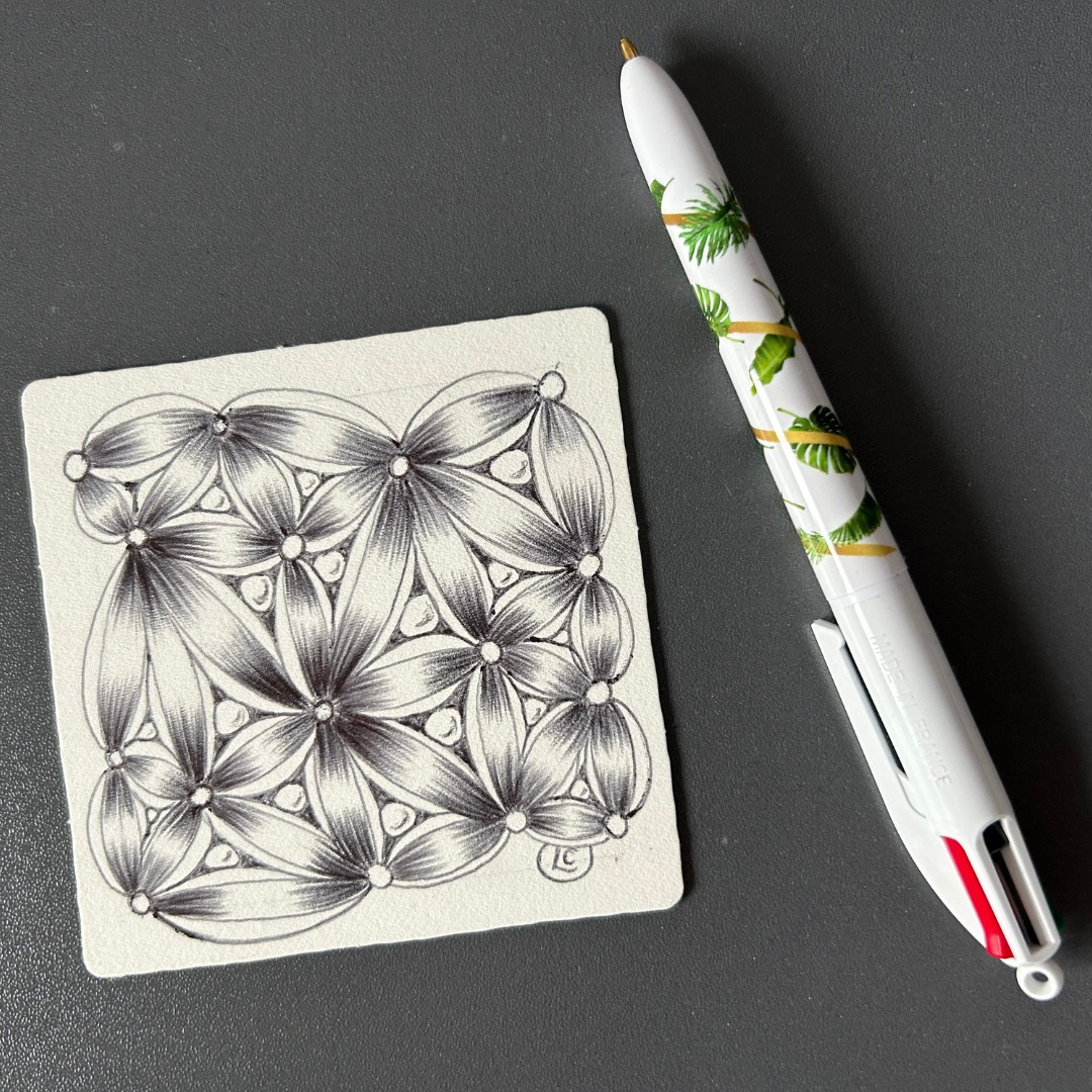 A paper tile that has tangles drawn on it using only a biro pen, which is sitting on the desk next to the tile. A paper tile that has tangles drawn on it using only a biro pen, which is sitting on the desk next to the tile.