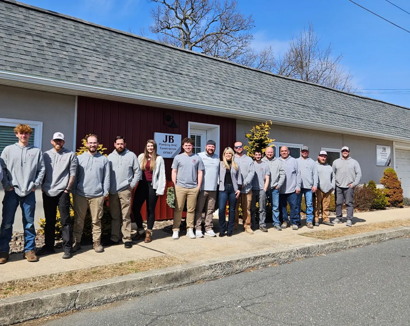The JB Plumbing HVAC & Construction team poses together in front of their office building, wearing matching branded gray shirts and hoodies. The group of approximately 15 employees stands outside the company's office, which displays a sign reading 'JB Plumbing HVAC Construction Office'. - JB Plumbing HVAC & Construction