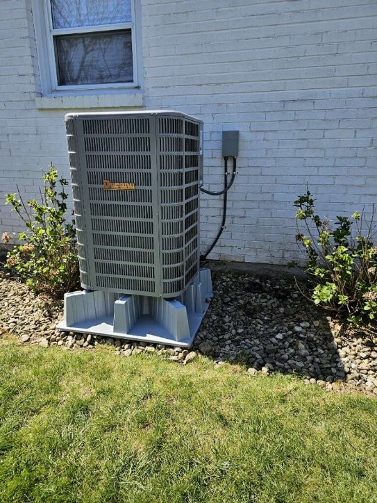 A newly installed Amana brand central air conditioning condenser unit mounted on a plastic pad base, positioned against a white brick exterior wall with proper electrical disconnect box and conduit wiring.