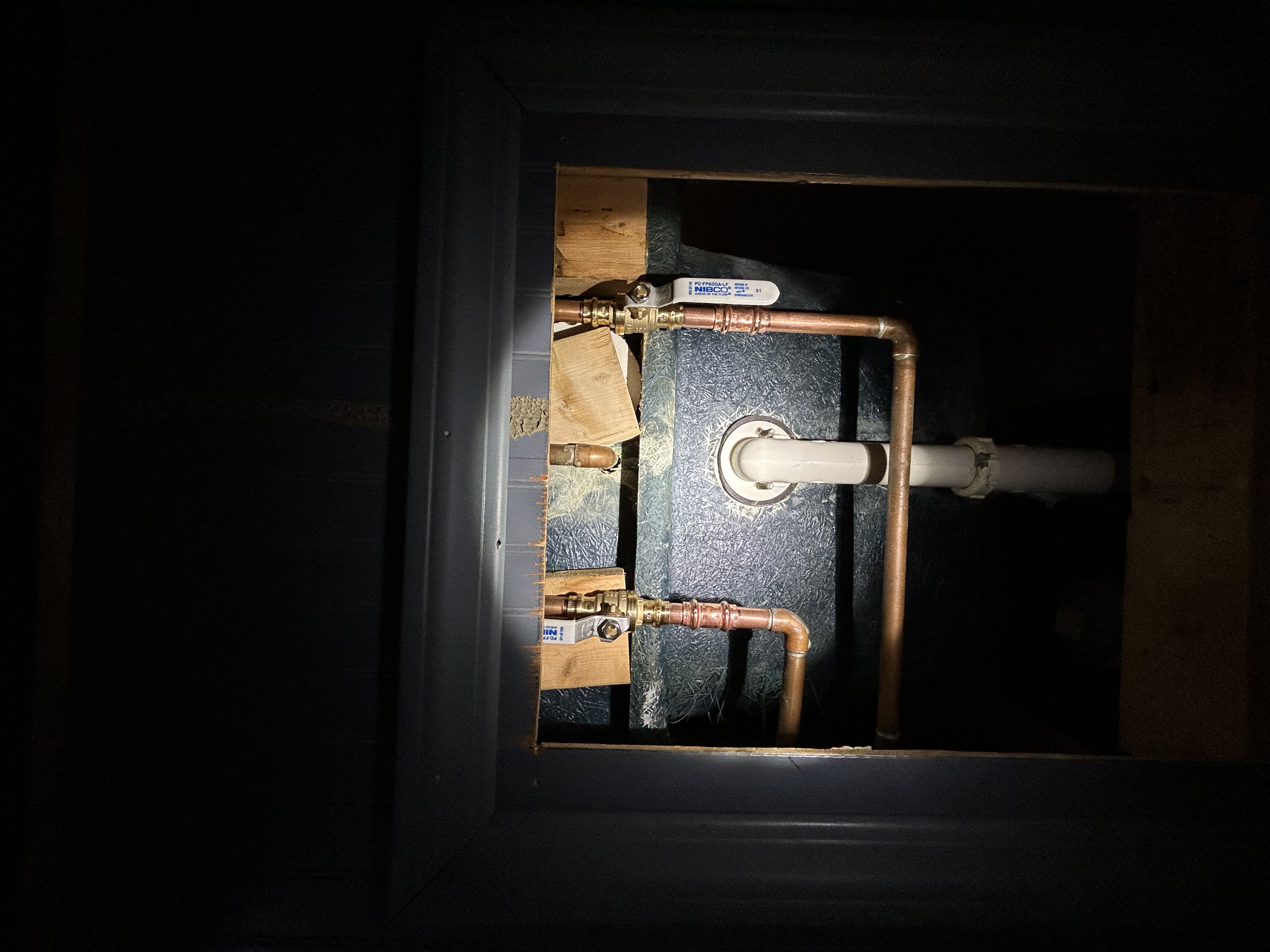 An access panel reveals copper supply lines with shut-off valves and a white PVC drain pipe, likely behind a bathtub or shower fixture, showing rough-in plumbing work with insulation board visible in the background.