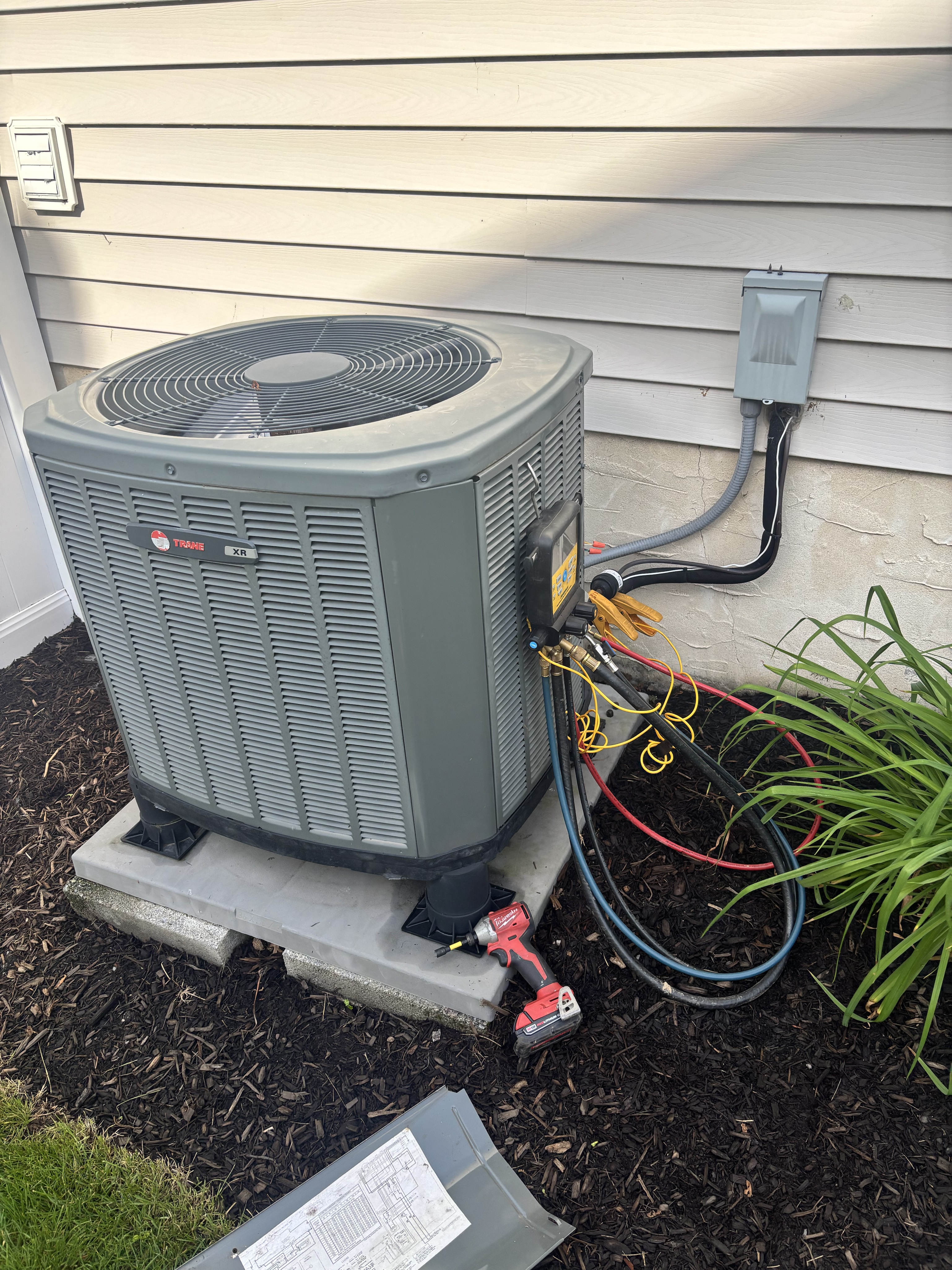 A Trane XR series outdoor AC condenser unit being serviced by an HVAC technician, with refrigerant manifold gauge hoses (blue, red and yellow) connected to the service ports during a refrigerant charge or diagnostic procedure.