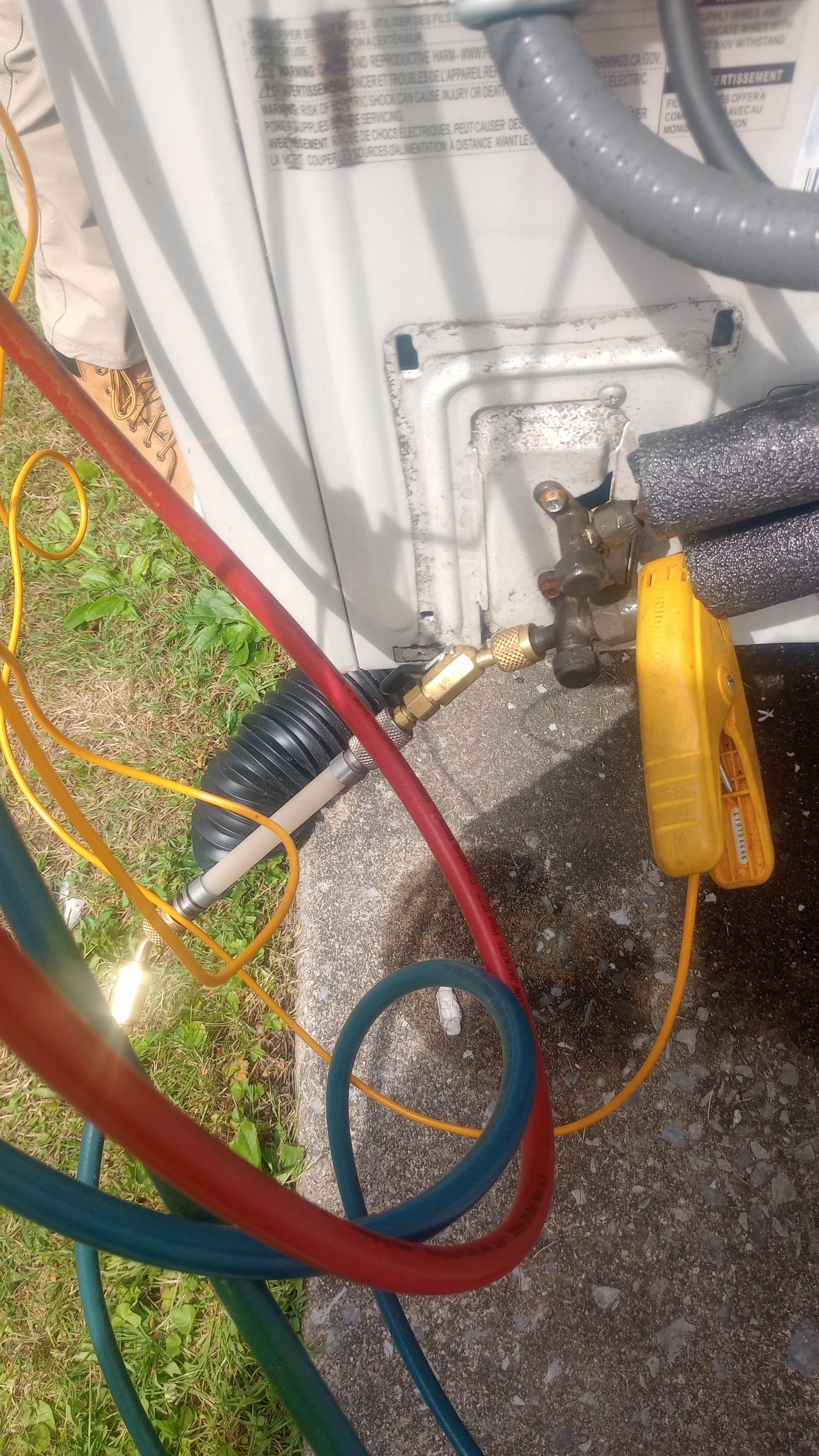An HVAC technician is servicing an outdoor AC condenser unit, with refrigerant manifold gauge hoses (red, blue, yellow) connected to the service valves, along with a refrigerant leak detector device visible on the right.