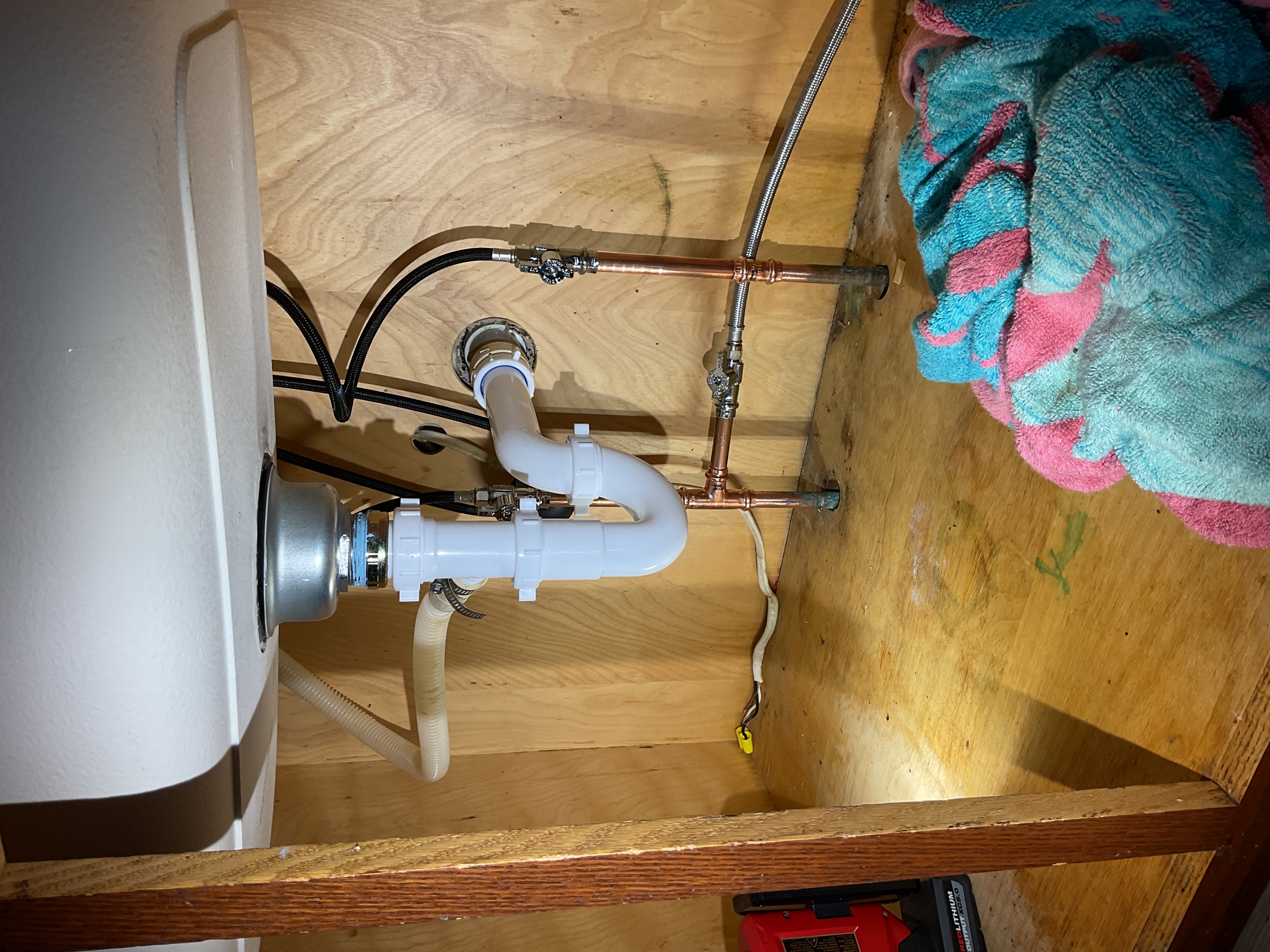 Under-sink plumbing installation showing a white PVC P-trap drain assembly connected to copper supply lines with shut-off valves, braided stainless steel supply hoses and black braided flexible lines inside a wooden cabinet.