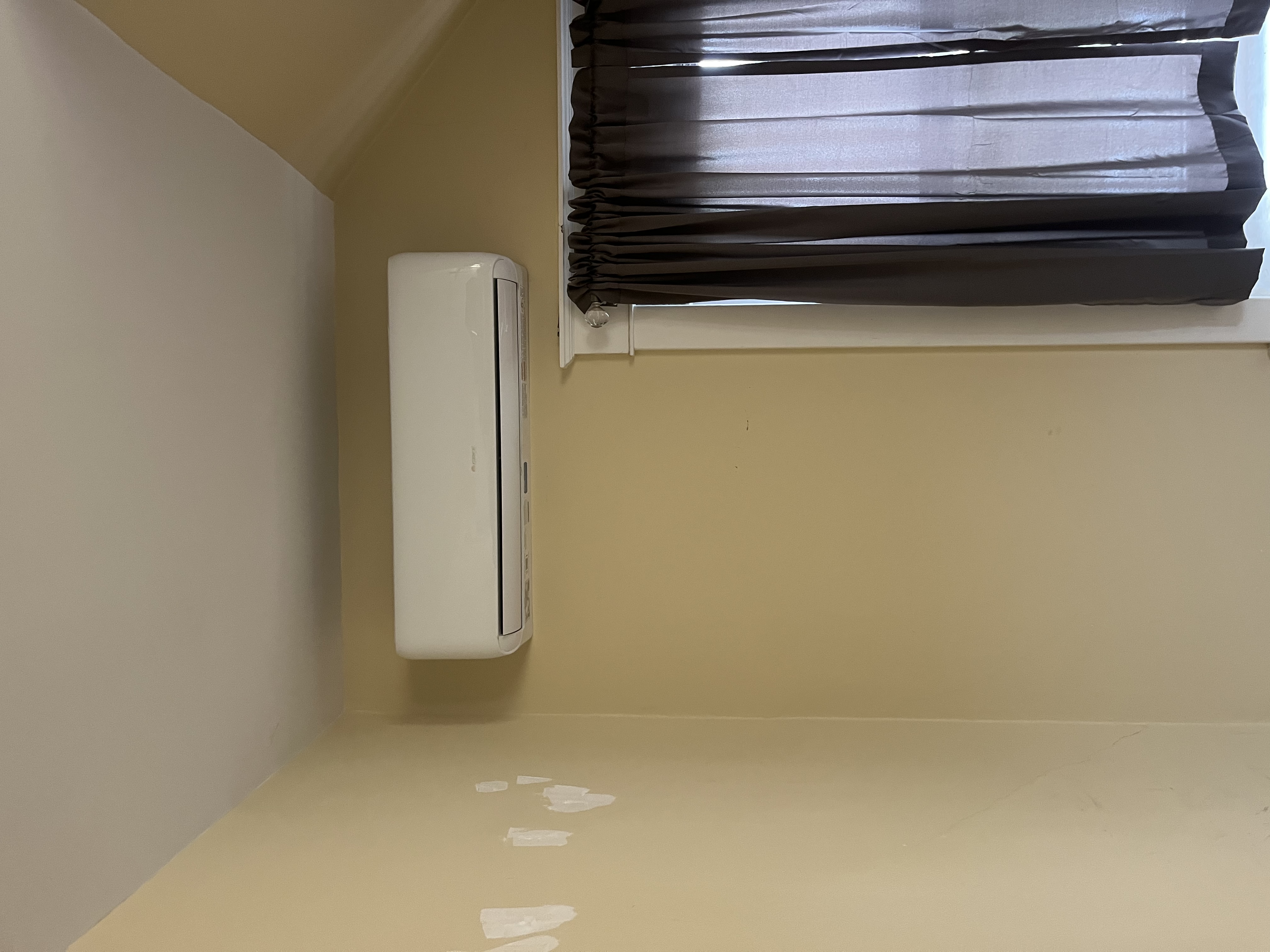 Ductless mini split head mounted high on a yellow wall beside a window.