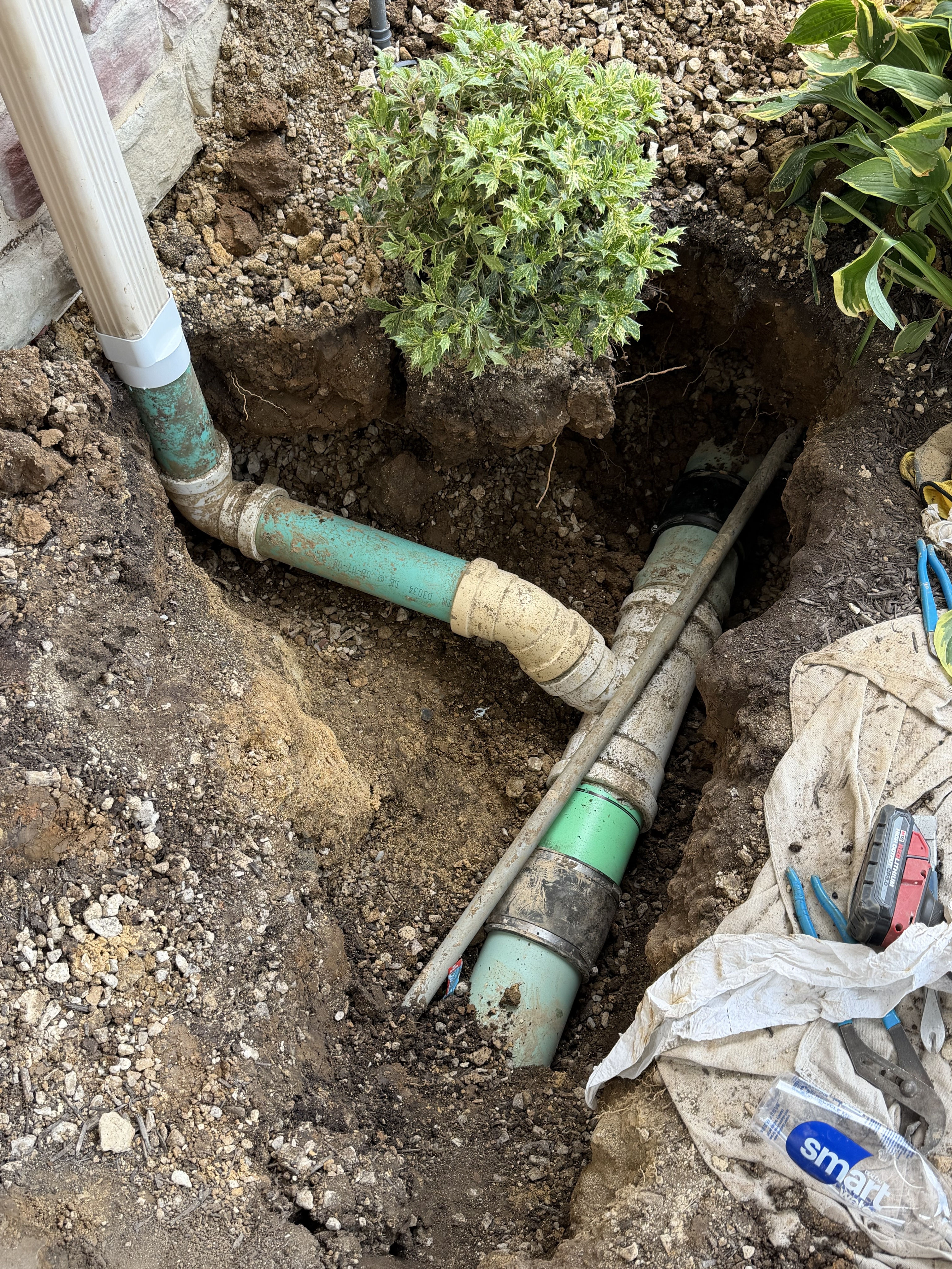 An excavated trench beside a house foundation revealing an underground drain/sewer pipe system with green ABS or PVC pipes, fittings and couplings being repaired or replaced, with plumbing tools visible on the side.