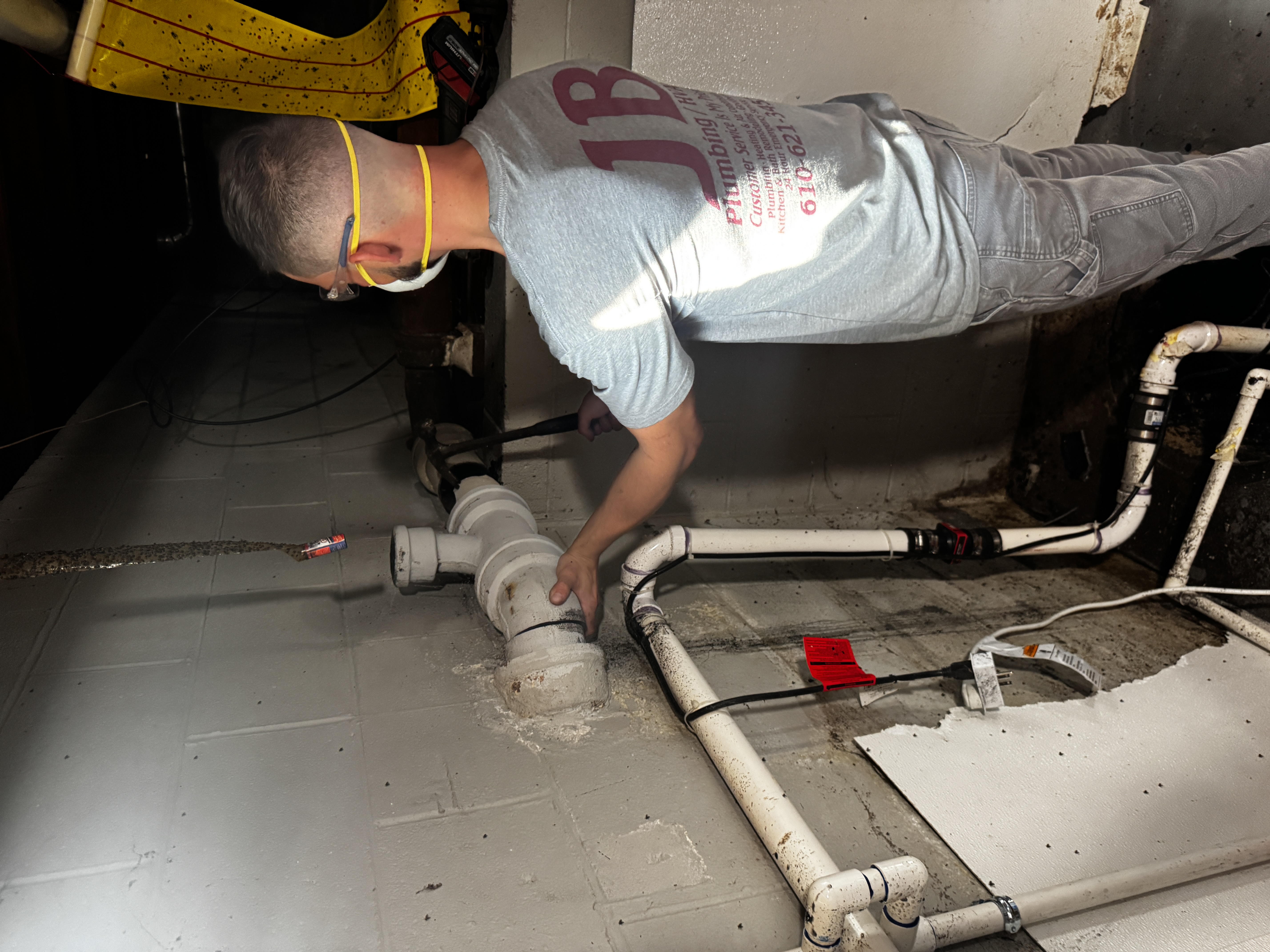 A JB Plumbing HVAC technician wearing safety glasses and a respirator mask inspects or installs PVC drain pipe fittings in a basement utility area, with multiple white pipes running along a concrete block wall.