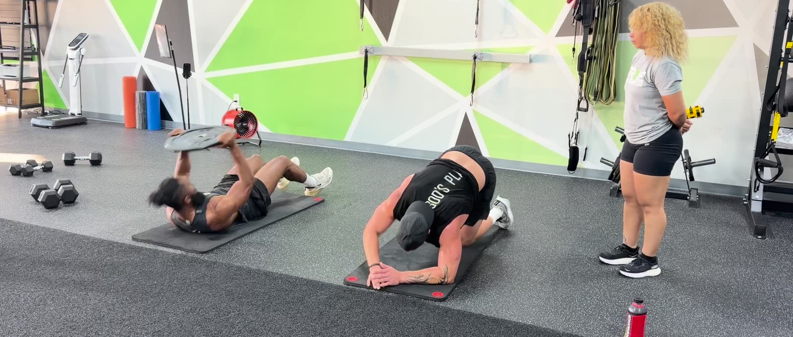 Two members working floor stations on mats beside the rack.