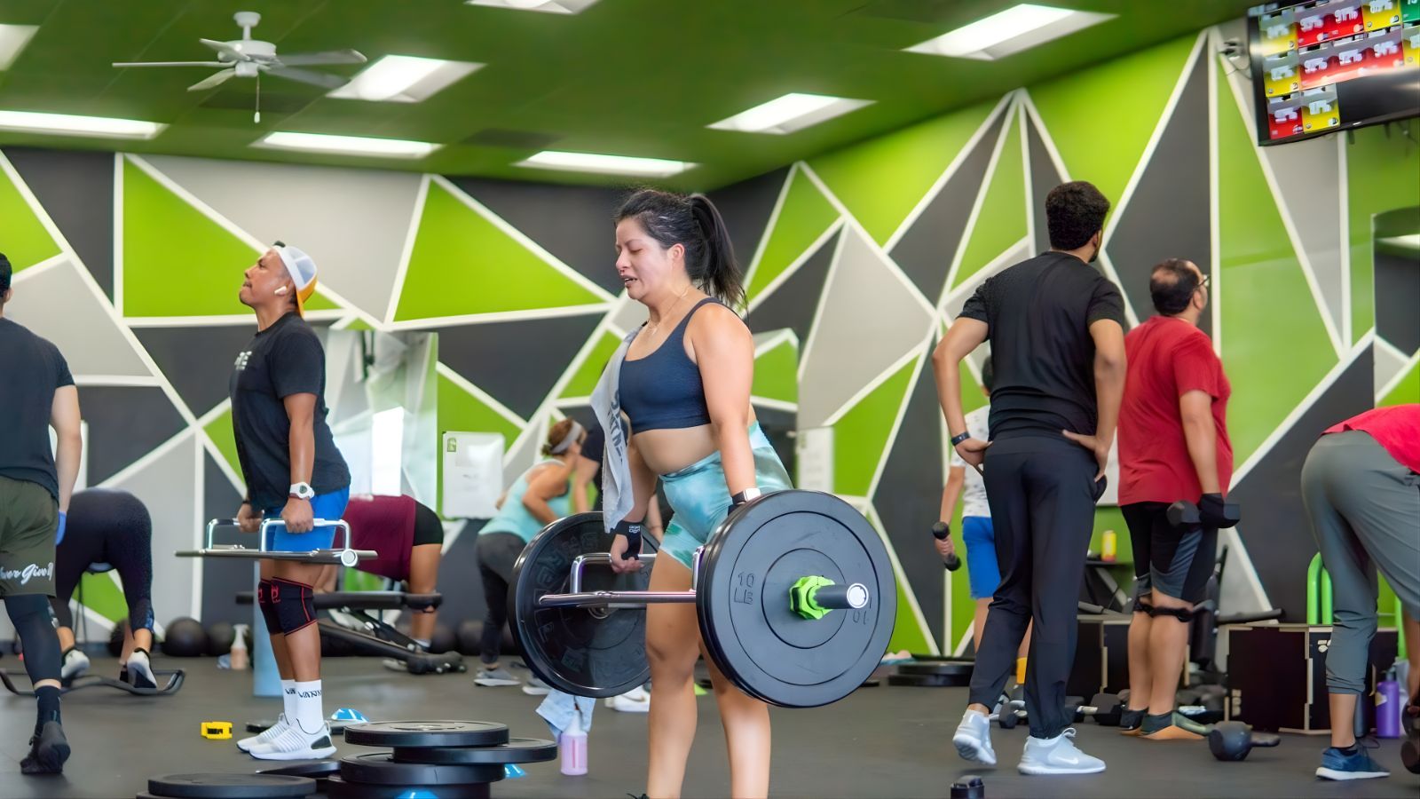 The floor of a Strive 11 Fitness gym: six members training at once on the open floor, each on a different movement, under the brand's green geometric wall.